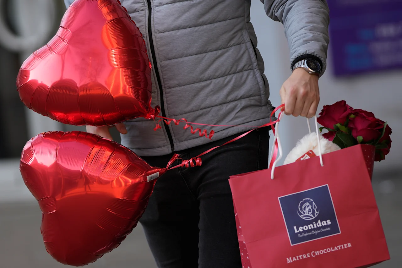 A man carries a bag with flowers and chocolates while holding onto two red balloons on Valentine's Day in London, Tuesday, Feb. 14, 2023. The economy gains from Valentin's Day as people in Britain spend around 1.37 billion pounds in 2022, that was was on flowers, food, entertainment and travel with some 25 million cards bought for the day. (AP Photo/Alastair Grant)