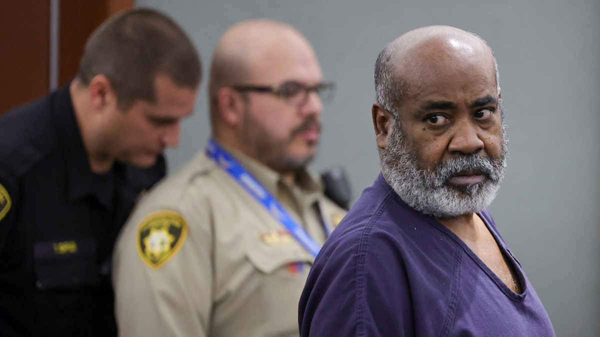 Davis, wearing a purple prison shirt, has a thick beard. He is looking intensely at someone or something off camera. In the background are two court law enforcement officers.