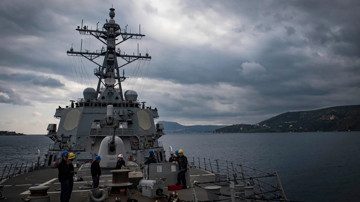 The ship pictured with land ahead, cloudy skies above. Personnel in hard hats stand on the ship.